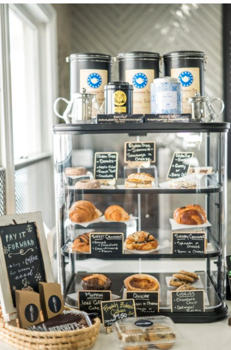 A display case filled with various pastries, quiches, and cookies, each labeled with chalkboard signs, sitting beneath coffee containers on top, with a 'Pay it Forward' board and a basket of coffee sleeves to the side